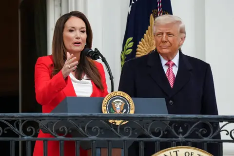 EPA/Shutterstock US Secretary of Agriculture Brooke Rollins wears a red blazer and peaks as President Donald Trump listens during an event celebrating farmers and Agriculture Day on the South Lawn of the White House in Washington, DC, on 27 March 2026.