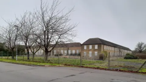Brick building with multiple boarded up windows. There are trees and grass in front of it.