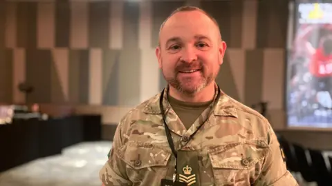 A man in army camouflage uniform smiles at the camera.