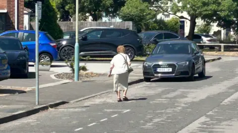 Picture of a street, cars are in the driveways of the homes and an older lady is walking on the road as opposed to the pavement. A car is also in front of her. She's wearing a white jacket and long dress. There's trees in the background of the image.