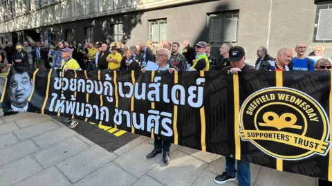 Oli Constable/BBC Around 60 supporters stand behind a black and yellow banner, with a message written on it in Thai in white lettering. Chansiri's face is on the side of the banner.