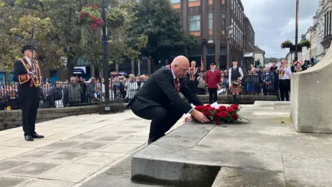 A man laying a wreath at the cenotaph in the Diamond, Londonderry, while a crowd looks on.