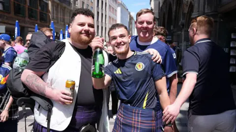 PA Med Craig Ferguson in the centre with two friends. He is wearing a Scotland football shirt which is dark blue with yellow and white flashes and a dark blue kilt with red, white and green flashes. He is holding a green bottle.