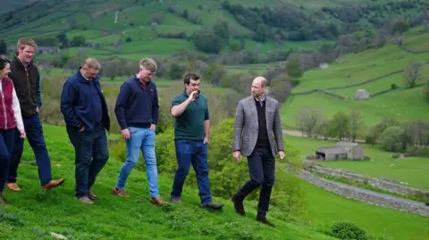Owen Humphreys/PA The Prince of Wales walks in a line with a group of five farmers. The scenery in the background is hills and lush green fields. He is wearing a grey suit jacket and has a hand on his hip. The farmers are mostly wearing blue jeans and brown or green quarter zip jumpers.