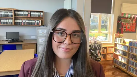 Sophie, a teenage girl with dark hair and large black rimmed glasses. She is wearing a claret blazer and a blue and white striped blouse. There is a brown table in the background and bookcases to the right.