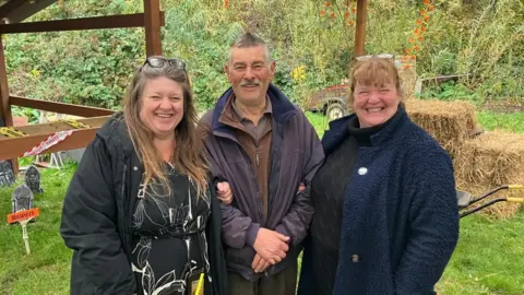 Three people: two women and one man pose on a farm - behind them are Halloween decorations and hay bales. The woman on the left has long brown hair with glasses on her head and is wearing a black floral dress. The man in the middle has short grey hair, a grey moustache and is wearing a short blue coat over a brown fleece jumper and the woman on the right has a beaming smile, is wearing a blue coat over a black jumper and has ginger hair.