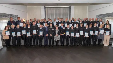 The attendees of the ceremony gathered for a group photograph. They are stood in lines behind one another. Many of them are in their uniforms and holding certificates. 