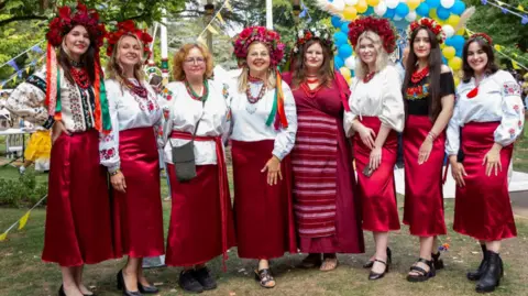Swindon Welcomes Ukraine Eight women standing standing in a line in a park. They are all wearing traditional Ukrainian dress - red skirts and white embroidered blouses and floral crowns. There are trees in the background and a huge bunch of white, blue and yellow balloons.