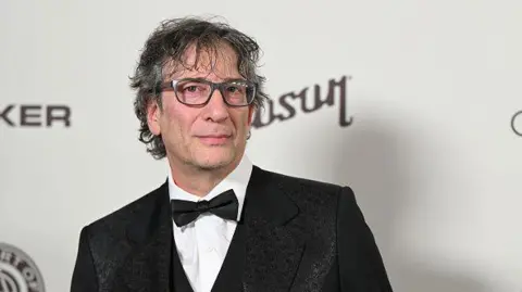 Getty Image shows Neil Gaiman at The Art Of Elysium's 2024 HEAVEN Gala at The Wiltern in Los Angeles, California, US, on 6 January 2024. He is wearing a suit and bow tie and is looking at the camera 