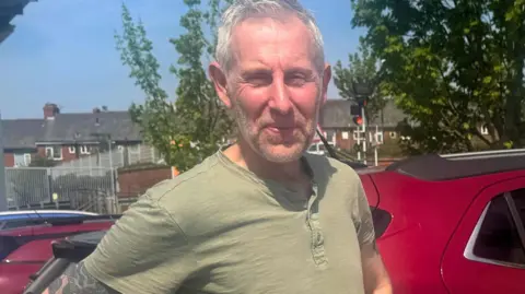 BBC/ Emily Johnson A man with short grey hair and a khaki t-shirt stands in a car park.
