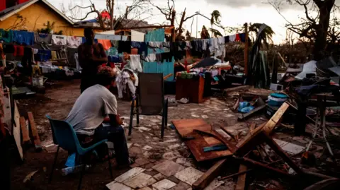 Reuters A man sits on a chair behind a destroyed house in the wake of Cyclone Chido as clouds appear overhead.