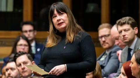 Anneliese Midgley has long brown hair and is wearing a black sweater. She is delivering a speech in the House of Commons as fellow MPs look on.