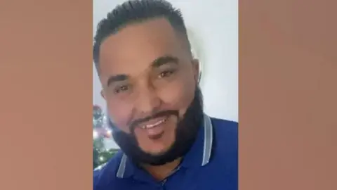Picture of Bonallie. He is tanned with short black hair and a thick black beard, wearing a blue polo shirt with part of a Christmas tree behind him
