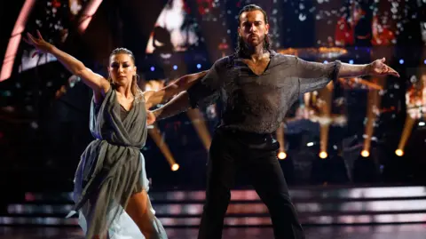 Jowita Przystał in a grey dress and Pete Wicks in a grey T-shirt and black trousers with their arms spread during their dance. Floor spotlights are visible in the background, behind grey steps.