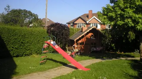 A red slide with a doll at the top of the slide and a wooden playhouse in the corner of the garden.