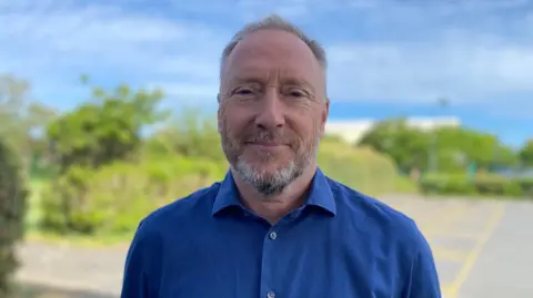 Deputy Paul Montague stands outdoors on a paved road, wearing a blue buttoned shirt, with greenery and trees behind, under a cloudy sky, photographed in natural daylight with shallow depth of field.
