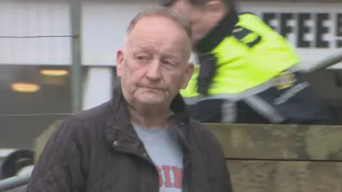 RTÉ Anthony McGinn stands in front of a wall, with two irish police officers in the background. He is wearing a grey t-shirt and a brown court. 