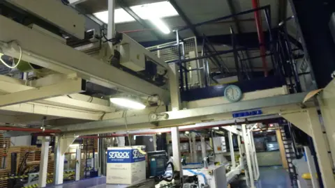 HSE A factory floor with machines overhead. There is a clock attached to a metal bar in the distance.