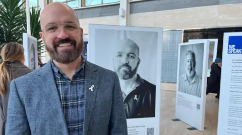 Nicola Haseler/BBC Mike Taggart also stands in front of the portrait of him that features in the exhibition. He is bald and wears glasses. He is wearing a blue-checked shirt underneath a grey jacket, which has a white ribbon pinned to the lapel. He is smiling. Other portraits in the exhibition can be seen behind him.