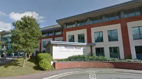 A Google Street View of Mid Devon District Council’s office. It is a brick and white coloured building. There is a outbuilding with the councils logo and the name of the building 'Phoenix House' on the side of it. Trees and grassland is visible.