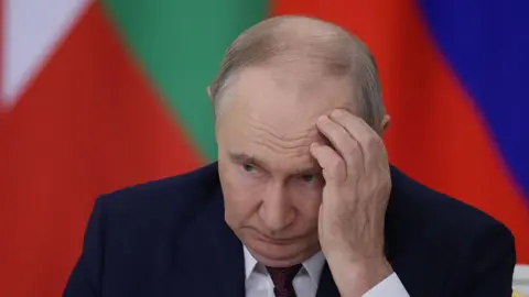 Getty Images Vladimir Putin sits hunched over in a dark blue suit and red spotted tie, with his left hand to his forehead. He looks tired and nervous. 