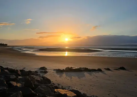 Moira MacKintosh A sunset over a beach. There are rocks in the foreground and the sky is yellow and blue. 