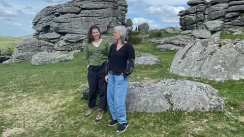 Two women stand in front of Hound Tor as they act out a folk story.