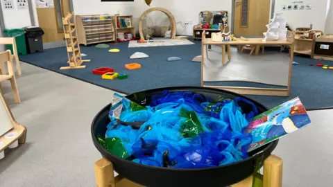 A large room is filled with toys suitable for young children who have sensory needs. There are large colourful wooden blocks and shapes on the blue carpeted floor, small wooden chairs around a wooden table, and a large dish in a wooden stand full of pieces of blue fabric and green plastic sheeting.