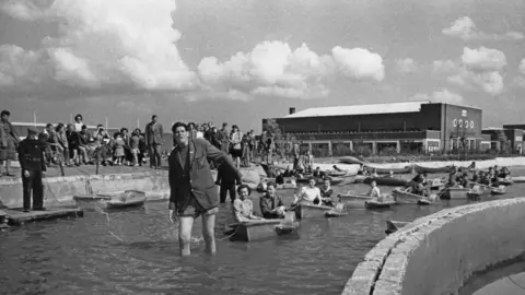 Getty Images/Charles Hewitt A black and white image of a holiday park in the 1940s with a man wading in water pulling a series of small boats with people in them