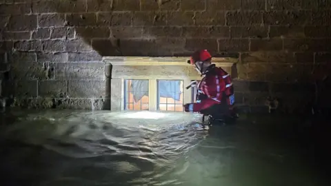 Northamptonshire Search and Rescue A member of Northamptonshire Search and Rescue wades through flood water. He is wearing red high-visibility clothing and a red hard hat. He is holding a torch while walking past a window, where the flood water is high enough to have covered part of it.