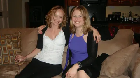 Family photo Two women sit on a brown sofa with their arms around each other. They are smiling at the camera. One is holding a glass of white wine in her right hand.