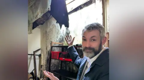 Woking MP Will Forster pictured inside his former music classroom which has now been retired from use. Will is holding his hands aloft while gesturing at the condition of the classroom. Debris can be seen on the floor with huge amounts of mold in the walls. 