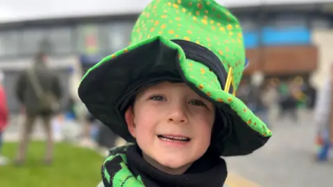 A boy is smiling. He's wearing a green leprechaun hat with a black buckle and gild strap. The hat is decorated with little gold flecks. The background is blurred. 