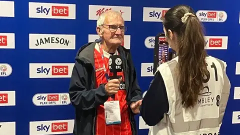 Gary Edwards Gary Edwards stands in the media area on his VIP tour of Wembley. He is holding an EFL microphone in front of a sponsor media board, and stood in front of woman who appears to be interviewing him. She has long brown hair and is wearing a white and cream bodywarmer that has Wembley written on the back.