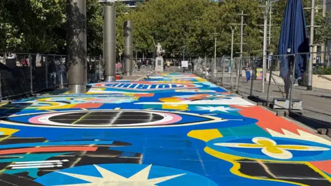 A large, bright and colourful mural painted on a street. It has lots of colours and shapes in it with the words 'Bristol' in a large heart.