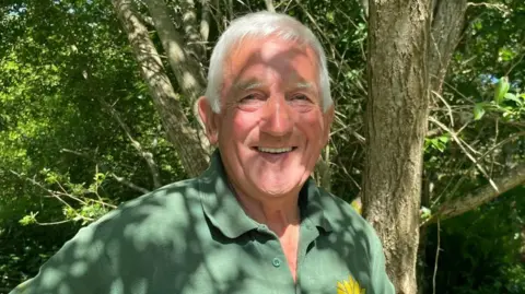 Norman Trebilcock smiles at the camera while he stands in front of a wooded area at Lanjeth Nursery and Water Gardens near St Austell in Cornwall. Shadow from tree branches is covering his face and chest. He is wearing a green polo shirt with a yellow flower on it.