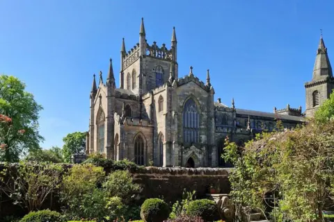 Nancy Chambers Dunfermline Abbey, an old light-coloured greyish church with several small spires. The surrounding gardens can be seen, with trees and bushes and a separating wall. The sky is blue.