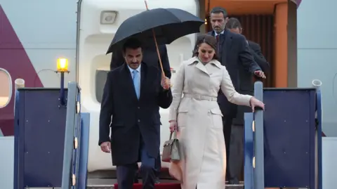 PA Media The Emir of Qatar Sheikh Tamim bin Hamad Al Thani, and Sheikha Jawaher, walk under an umbrella as they descent some steps from a plane arriving at Stansted Airport in Essex