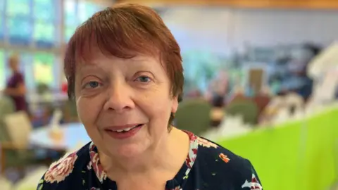 Eileen Wright, manager of Endike Community Care, looks directly at the camera. Behind her, blurred out, are clients sitting around circular tables. 