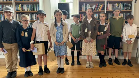 Ten children from the school all lined up for a picture. They are all dressed the fashion of the 1940s, and are each carrying boxes for gas masks, and their papers.