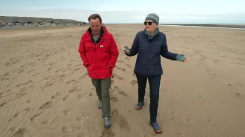 Wrapped up in wintry gloves David Braine is on the left of the shot as Liz talks to him about her love of photography. They are walking on a sandy beach and footprints can be seen near them.