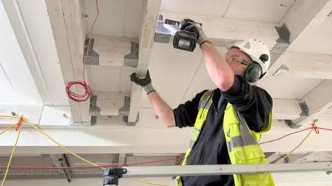 Builder with a hard hat, protective glasses and ear defenders using a hand drill to fix a beam in a ceiling. 