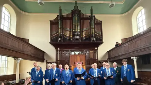 A male voice choir dressed in blue blazers and blue ties and holding choral hymn books sing in front of a large ornate pipe organ. 