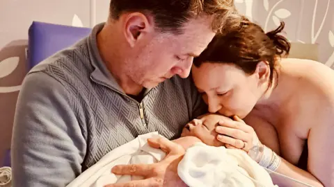 PA Media Rhodri Thomas holds baby Liliwen in his arms while his wife Emily Brazier kisses her forehead with one hand on her head.