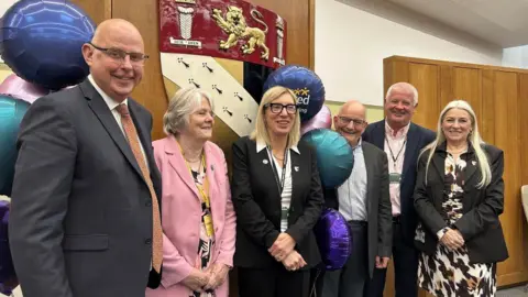 Kay Mason Billig/Norfolk Conservatives Three women and three men stand in front of the Norfolk County Council crest inside a building while smiling at the camera with balloons around them.