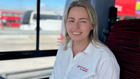 Kaleigh Hind is in the driving seat of a training bus at the Go North East depot in Gateshead. She is wearing a white shirt with Go North East branding and has shoulder length blonde hair. She is smiling widely and blurry red and white buses can be seen out the window to her side.