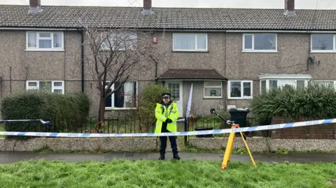 A policeman dressed in a yellow hi-vis jacket, black trousers and police cap standing outside the home where the incident occurred. It is a grey pebbledash two-storey home with a front garden and black railing gate. There is blue and white police cordon tape stretching across the front of the property.