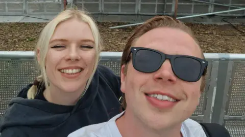A woman with blonde plaits wearing a black hoodie and a man with slicked back light brown hair wearing a white T shirt post for a selfie in front of a festival stage.