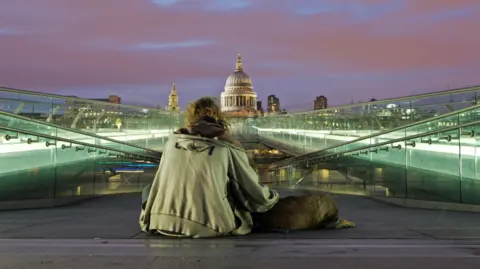 A homeless man sitting on the ground with his back to the camera, wearing a grey hoodie with a dog lying by his side. He is facing the dome of St Paul's Cathedral and on both sides of him are illuminated glass walkways