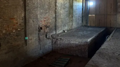 HAYLEY DOLBEAR The inside of a building which is made from brick. There is a bricked platform on the left which leaders towards a boarded up window which has a small square of light coming through.